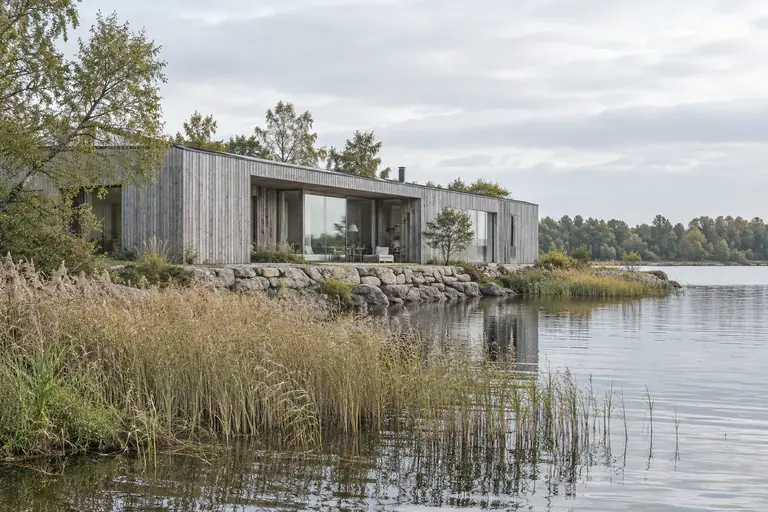 A low timber house beside a lake, framed by reeds and soft morning light.
